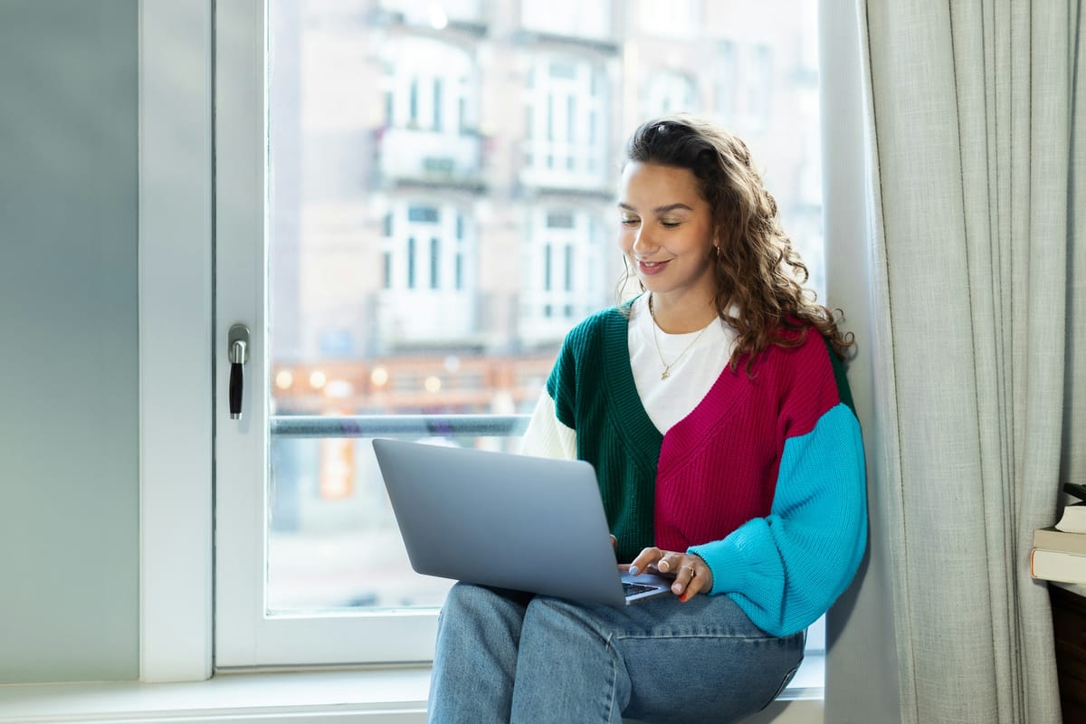 A woman sitting by a window at home with a laptop on her lap