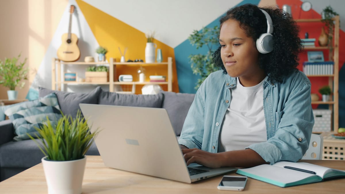 A student with headphones working on a laptop at a desk at home